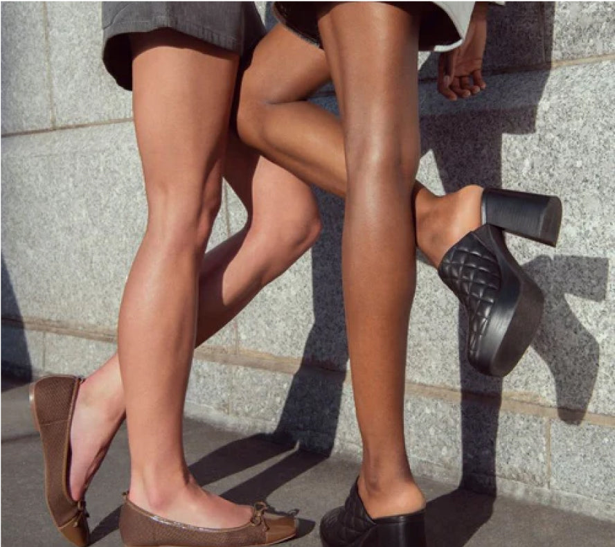 Two people standing, showing their legs and different shoes against a stone wall.
