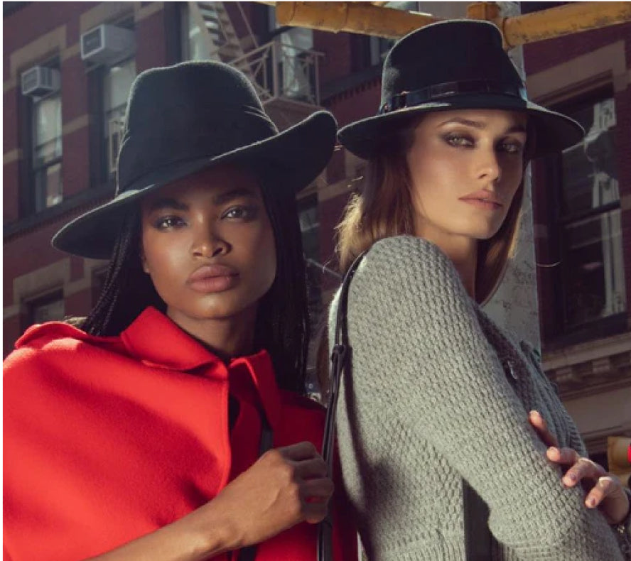 Two women in stylish coats and hats pose confidently on a city street.