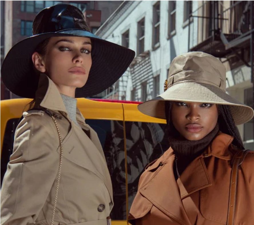 Two stylish women wearing wide-brimmed hats and trench coats in an urban setting.